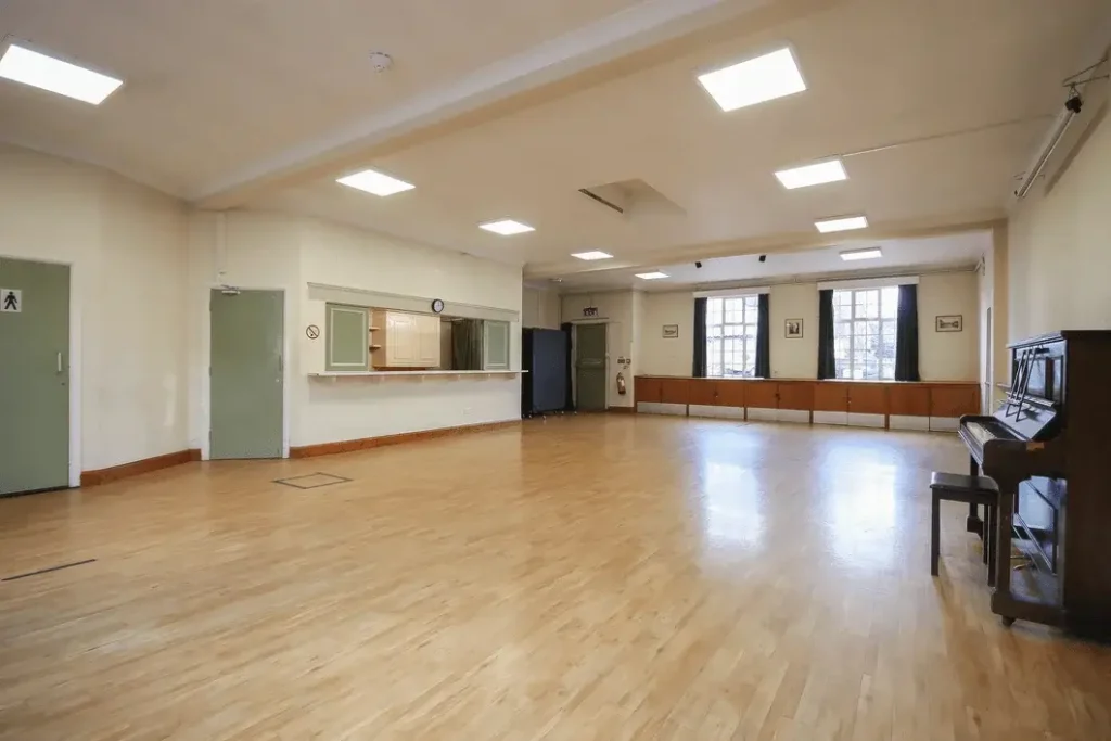 The Peter Griffits Hall is another venue within the Forest Row Village Hall. Image shows the interior of the hall, taken from the entrance towards the serving hatch, which is open. At the other side of the hall can be seen the emergency exit door.